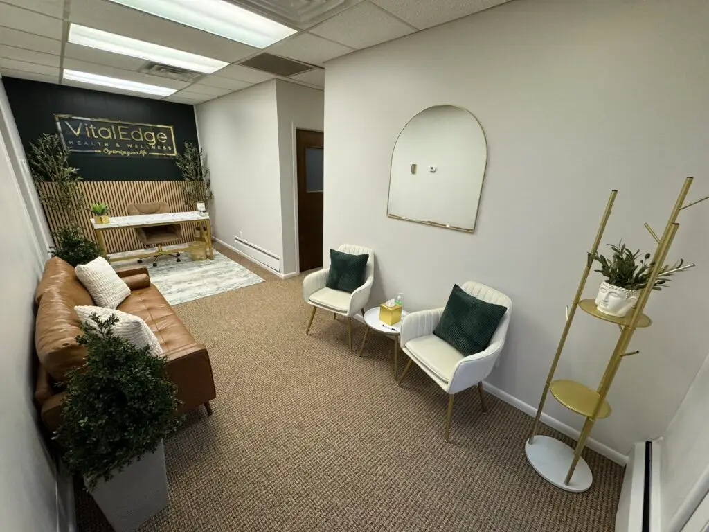 vitaledge health and wellness office with modern desk, brown leather sofa, white accent chairs, wall mirror, indoor plants, and gold shelving in a calm neutral-toned space