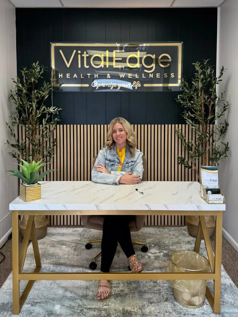adrienne s. o’neal fnp-c sitting at a marble desk in the vitaledge health and wellness office with branded sign plants and modern decor behind her