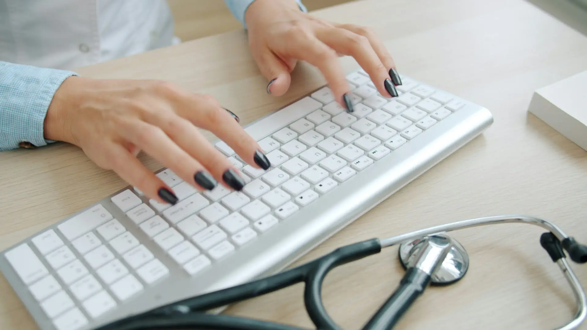 typing hands on a white keyboard with a stethoscope on the desk, representing medical documentation and patient care